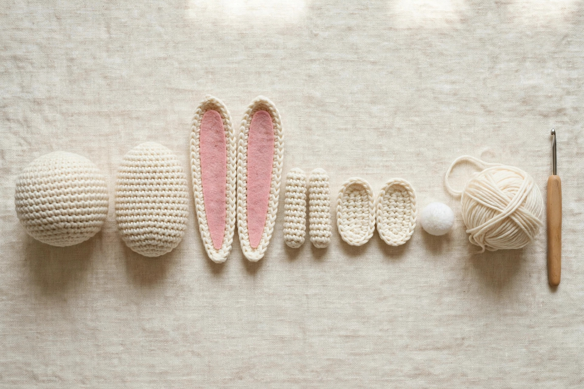 Crochet bunny pattern pieces laid out in a row, head, body, ears, arms, feet and pompom tail, next to yarn and a crochet hook