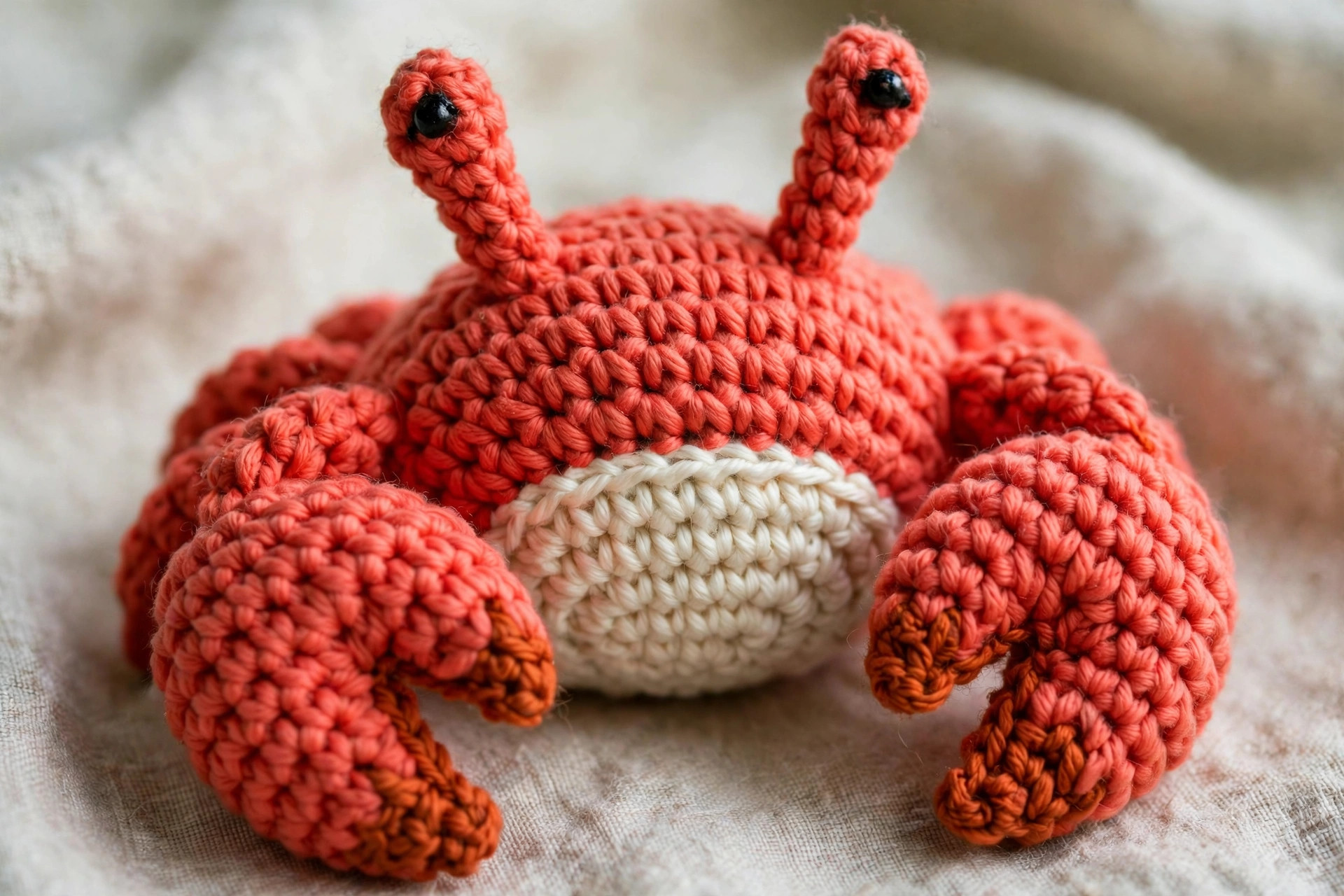 Close-up of a crochet crab showing the coral red body, cream belly, black eyes on stalks and orange claws