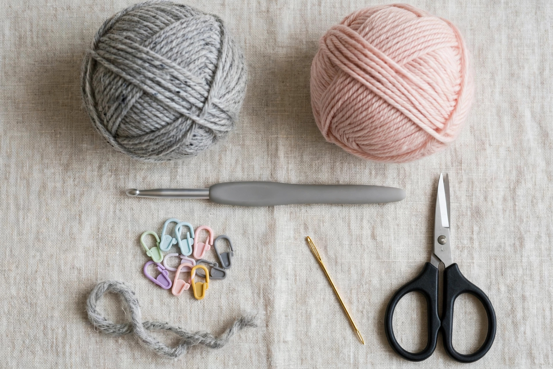 Flat lay of gray and pink yarn, a 5.0 mm crochet hook, tapestry needle, scissors and stitch markers for the crochet cat beanie pattern