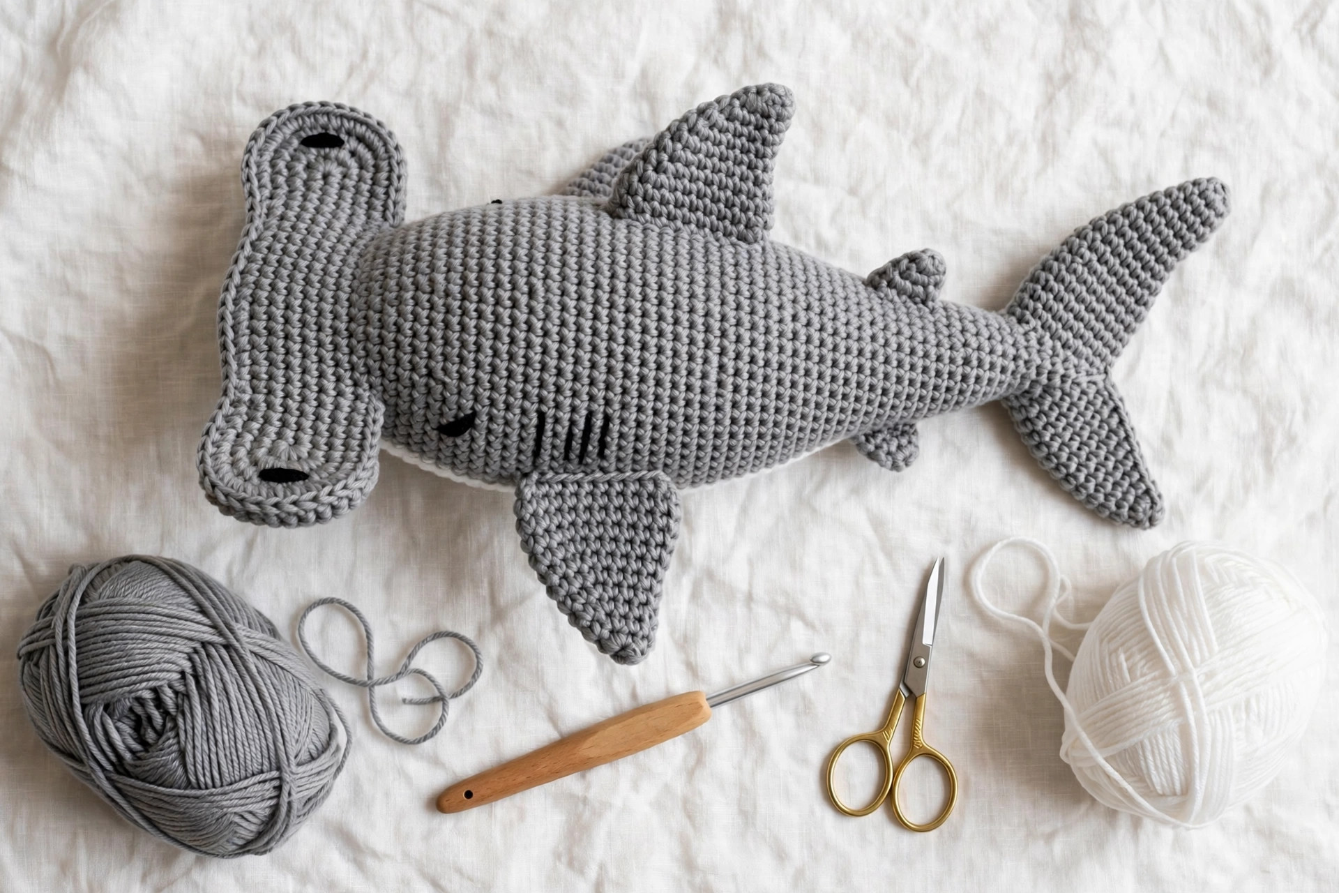 Flat lay of a finished grey crochet hammerhead shark with grey and white yarn balls, crochet hook and scissors