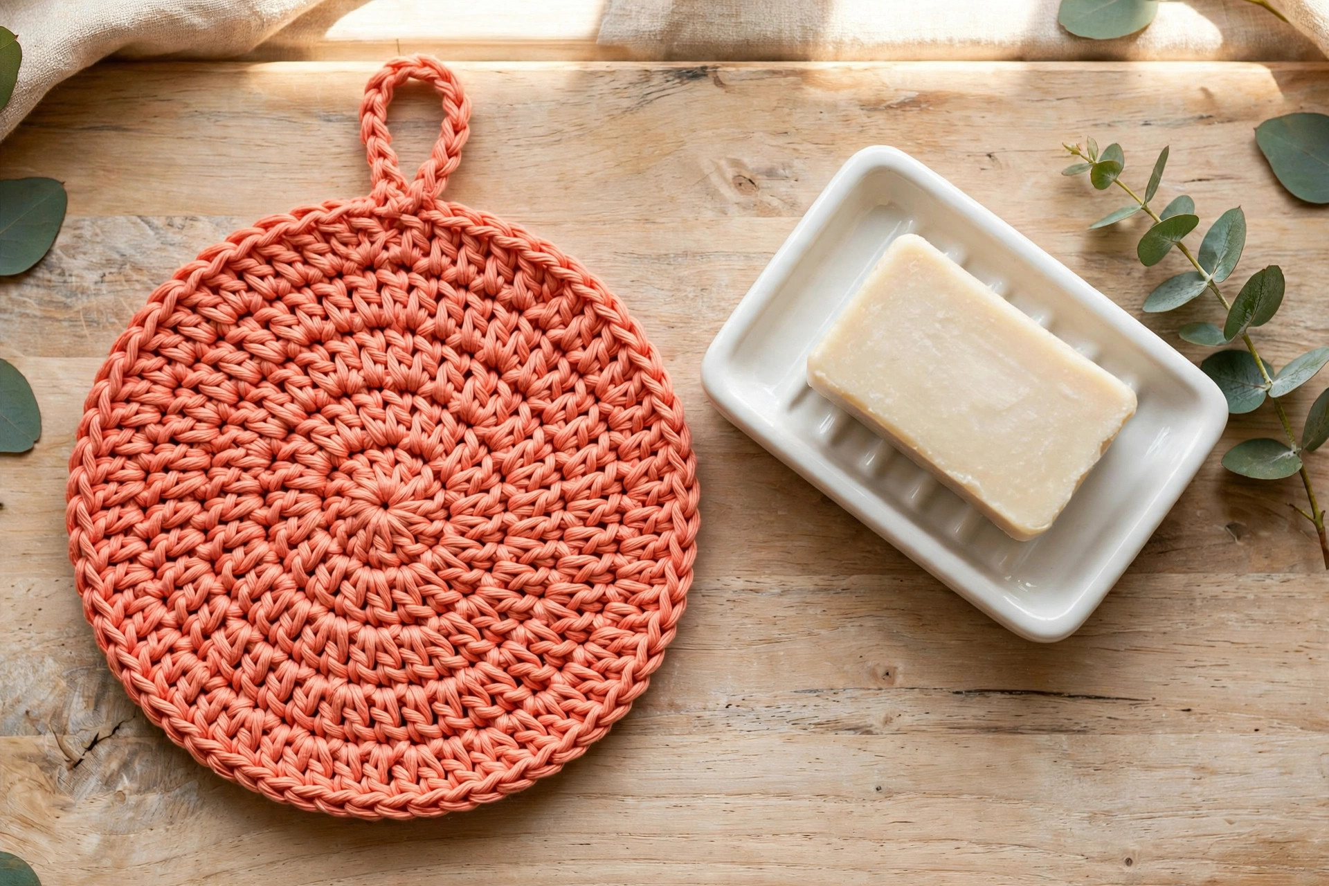 Coral round crochet scrubby dishcloth with a hanging loop next to a white ceramic soap dish on a wooden counter