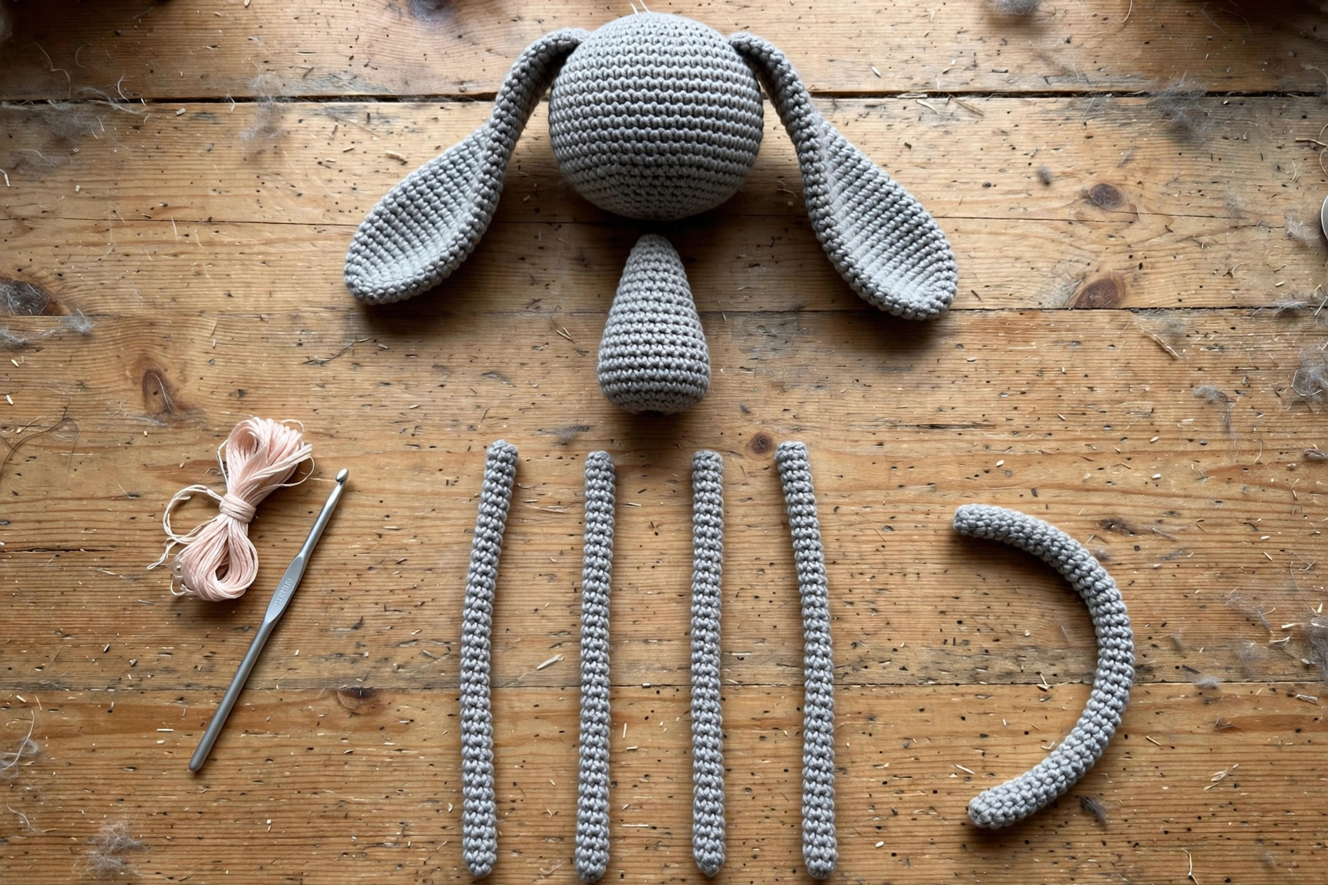 Flat lay of all crocheted floppy cat parts before assembly: round head, long floppy ears, tapered body, four long thin legs, curved tail, hook and yarn on a craft table