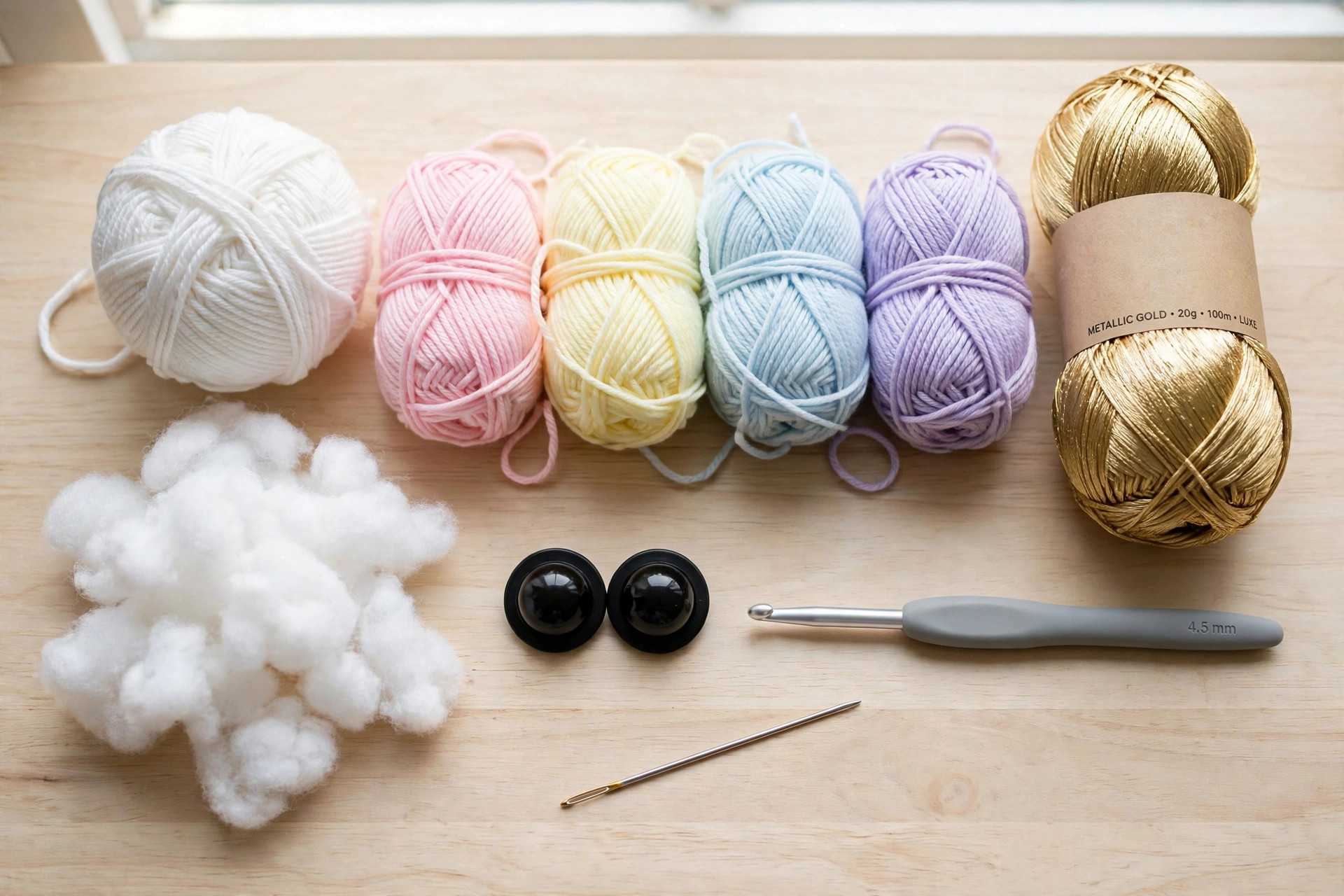 Flat lay of white and pastel rainbow yarn balls, metallic gold yarn, black safety eyes, a crochet hook, and toy stuffing on a wooden table, free crochet unicorn pattern materials