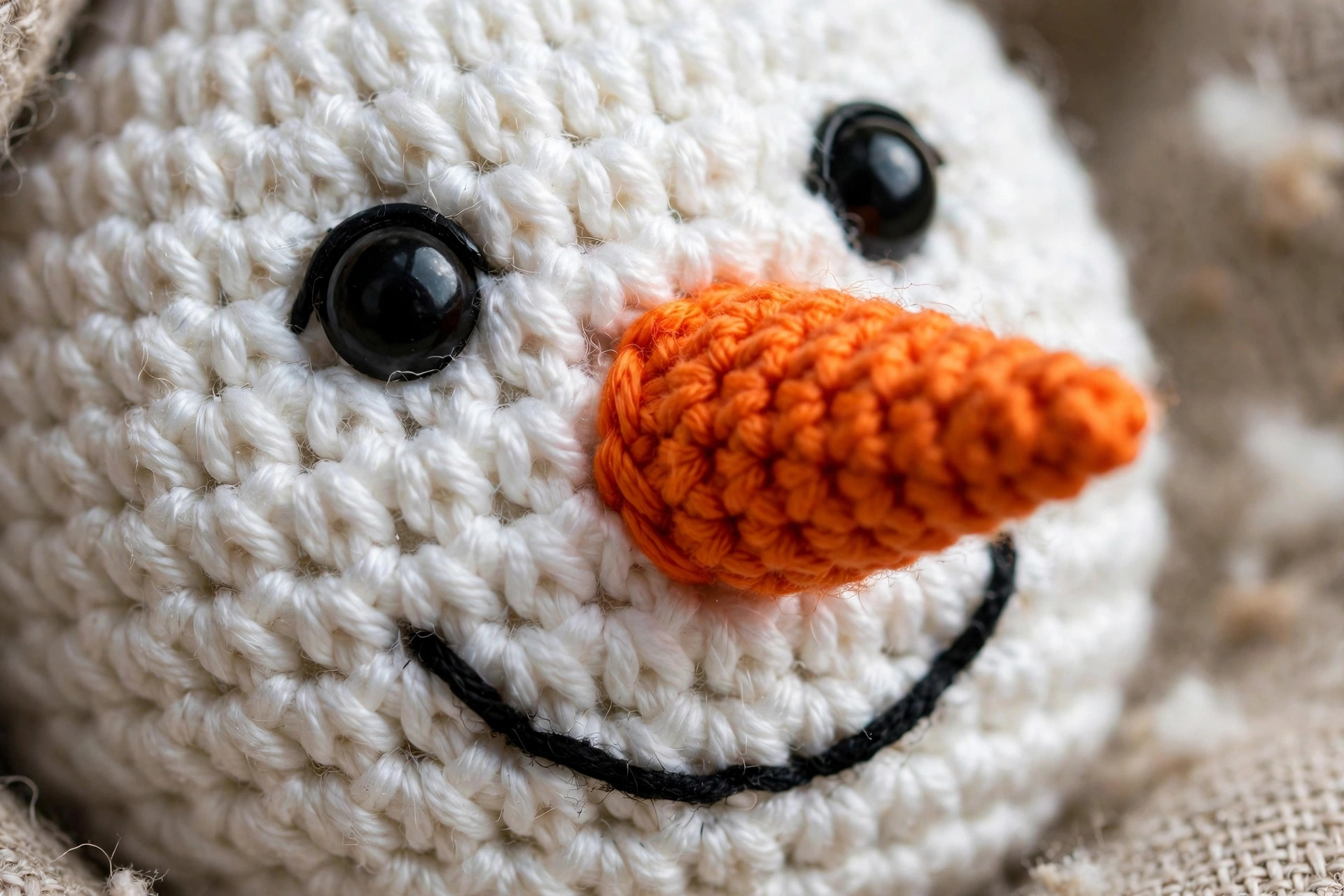 Close-up of a crochet snowman face with shiny black safety eyes, an orange cone carrot nose and a stitched black smile
